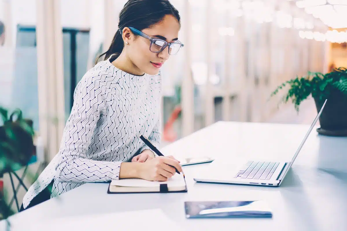 Une assistante administrative à lunettes écrit avec diligence dans un cahier à un bureau dans une entreprise.